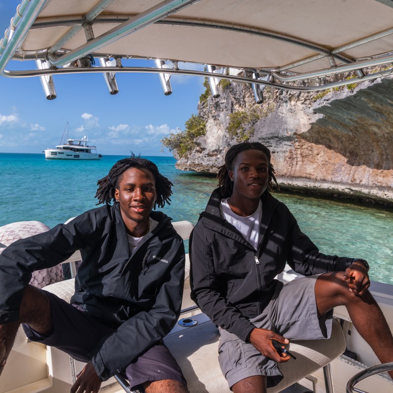 Two people on a boat near a rocky shoreline with a yacht in the background.