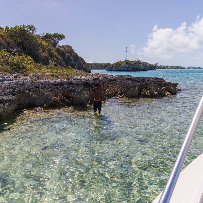 Person wading near rocky coast, clear blue water, boat in foreground, island background.