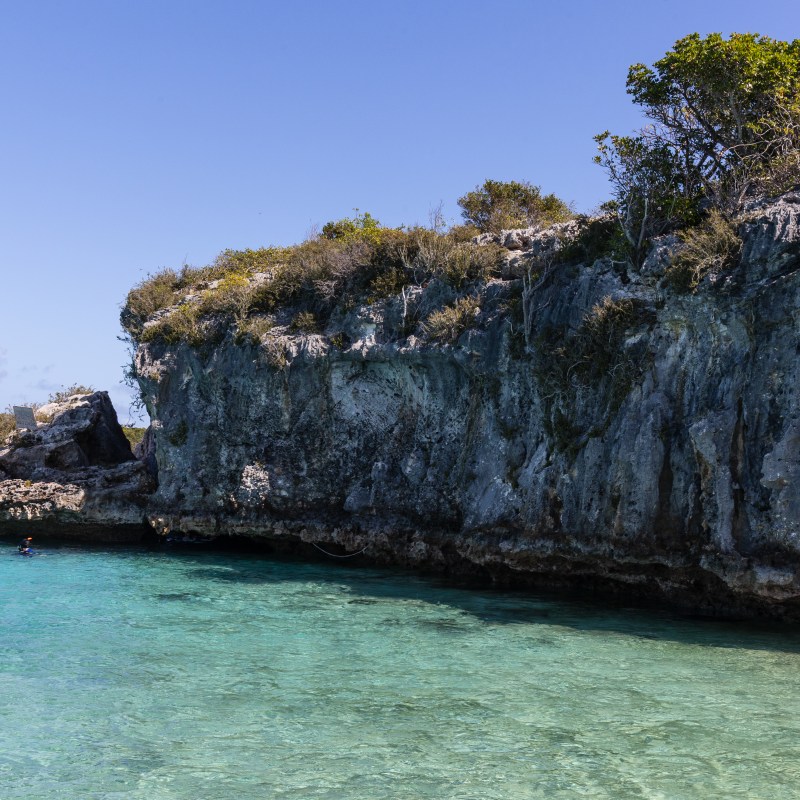 Cliffside above clear turquoise water with swimmers in the foreground.