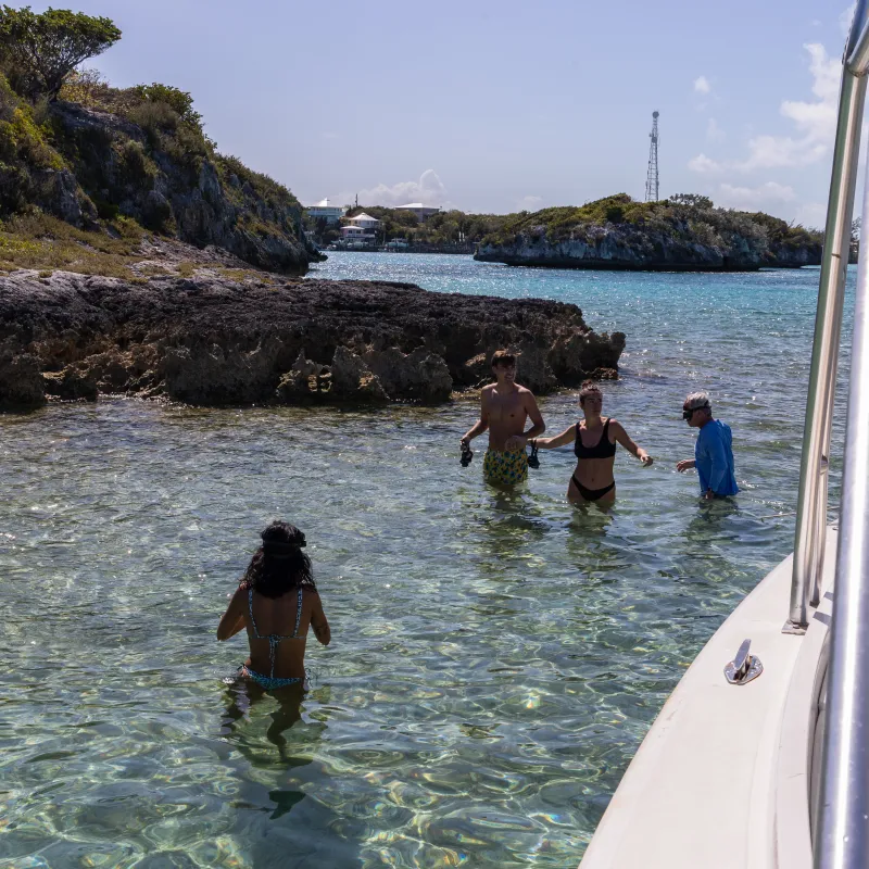 People wading in clear water near rocky shore, boat in foreground.