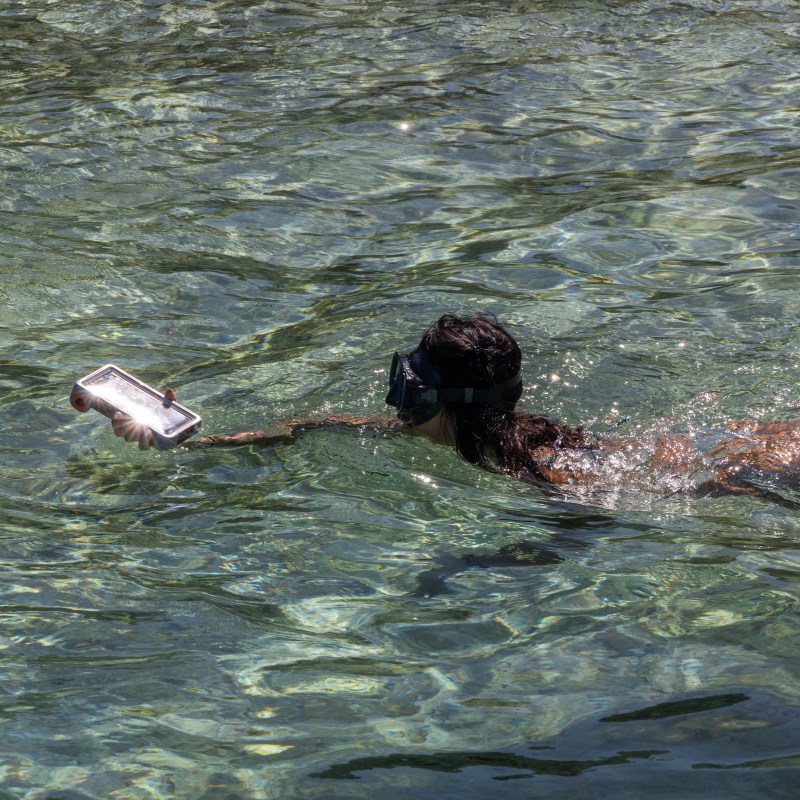 Person snorkeling in clear water holding a phone up.