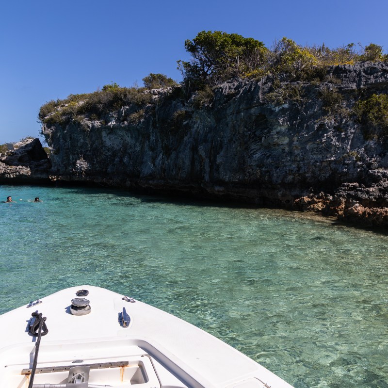 Boat approaching rocky shore with clear blue water and swimmers.