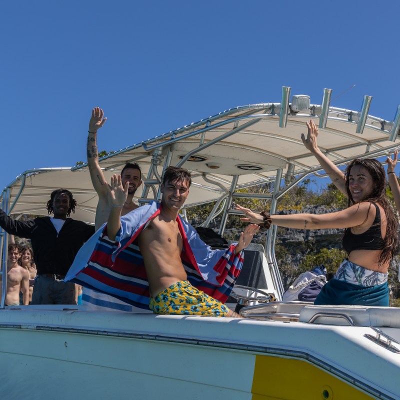 Group of people on a boat waving and smiling under a clear blue sky.