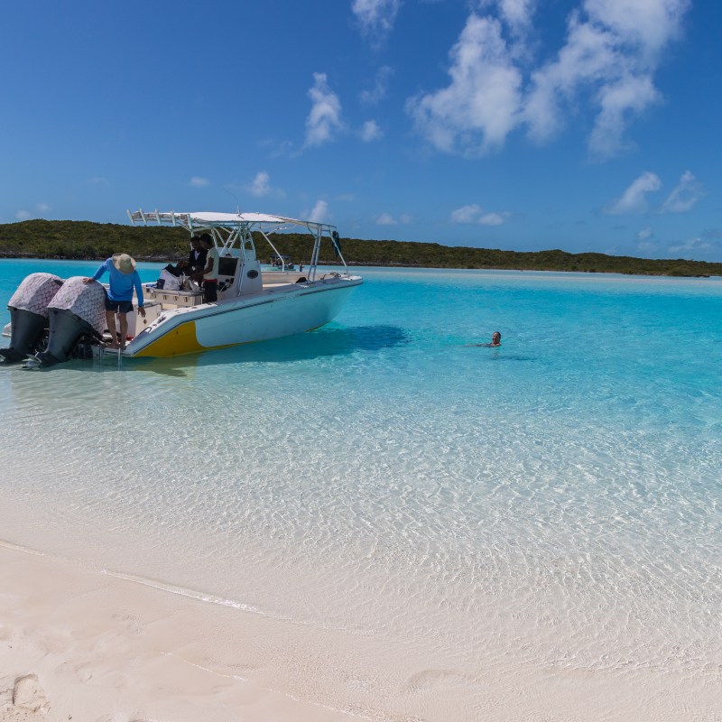 Boat anchored on clear blue water near a sandy beach with people nearby.