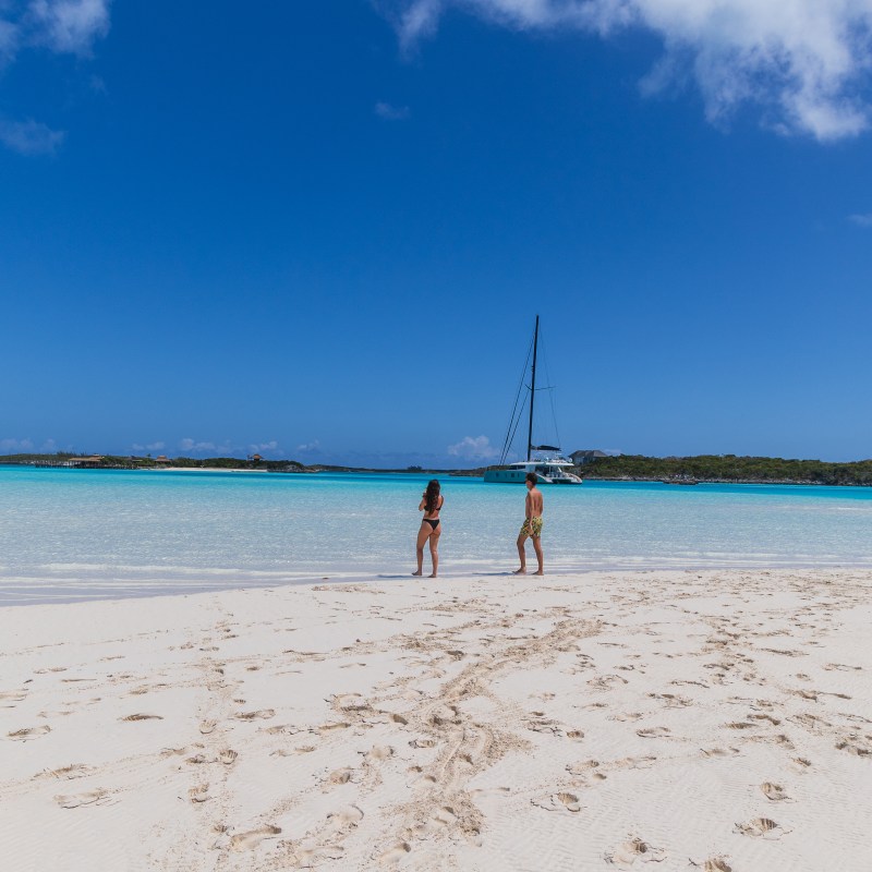 Two people walking on a sandy beach with a sailboat on turquoise water.