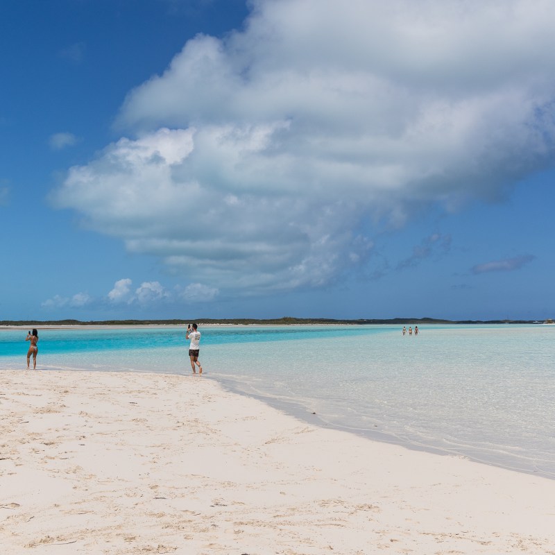 People walking on a sandy beach by turquoise water under a cloudy sky.