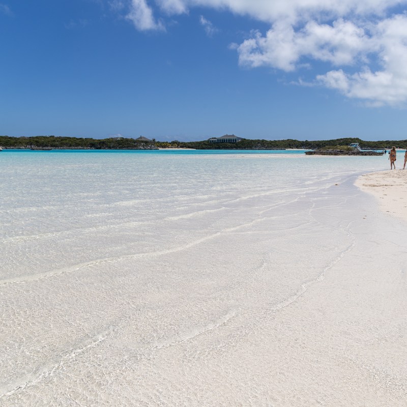Sandy beach with calm clear water, sailboat in distance, people walking along shoreline, sunny day with clouds.