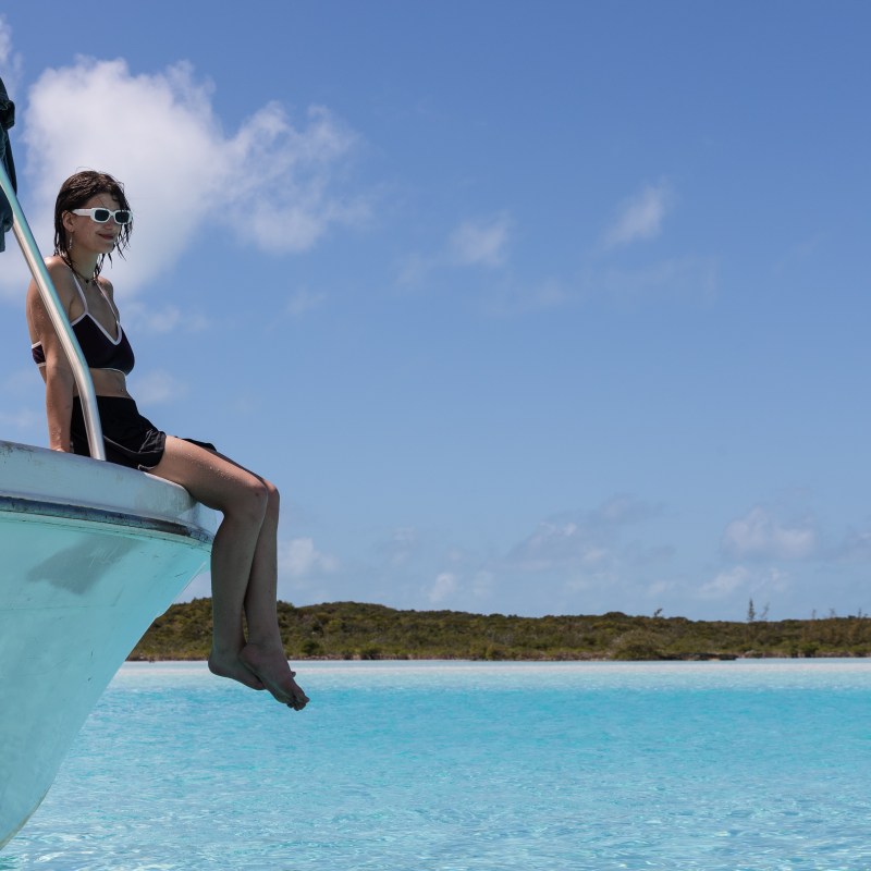 Woman sitting on boat edge, wearing sunglasses, with turquoise water and blue sky in background.