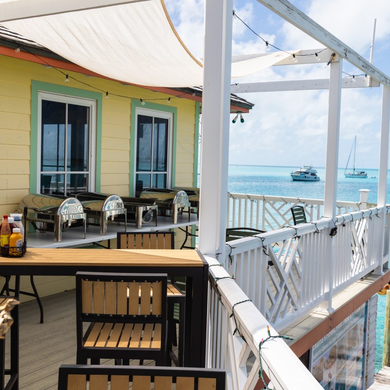 Seaside restaurant with patio seating, boats on turquoise water, and clear sky.